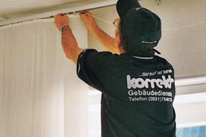 Mann in Korrekt-Arbeitskleidung montiert eine Vorhangschiene an der Decke.